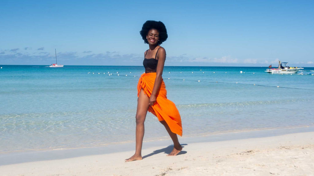 Woman smiling walking on the beach wearing an essential solid sarong by SHU-SHI in Orange